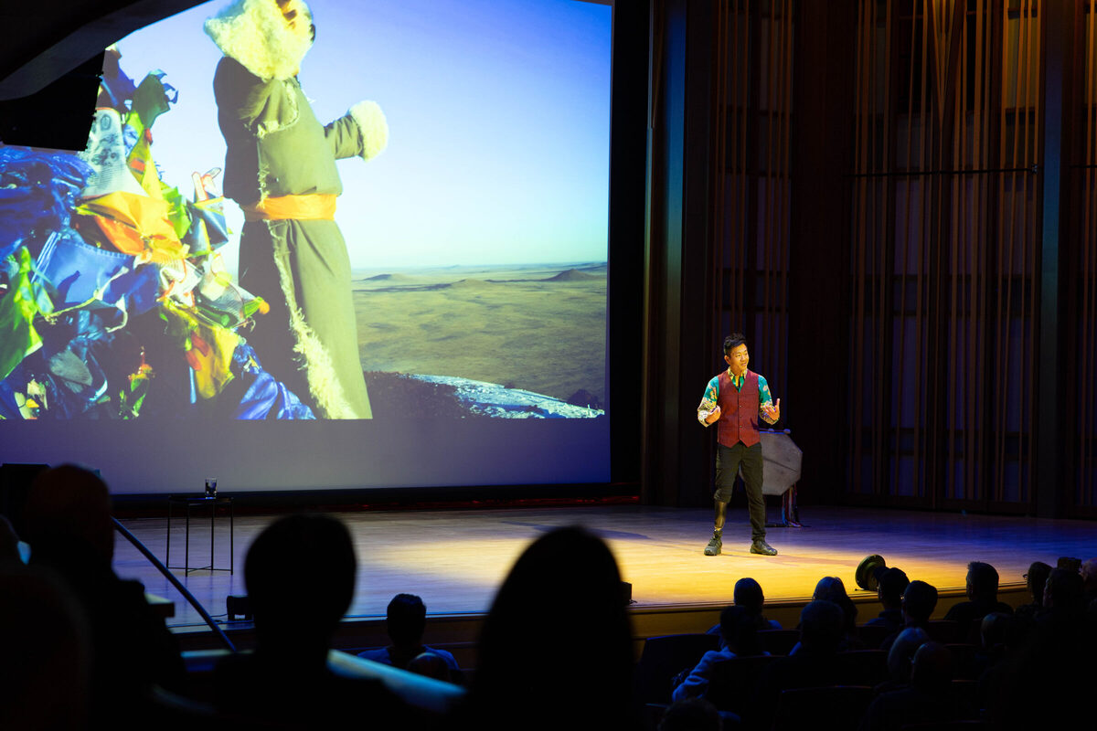 Albert Lin on a darkened stage before a seated audience, a vast landscape projected behind him